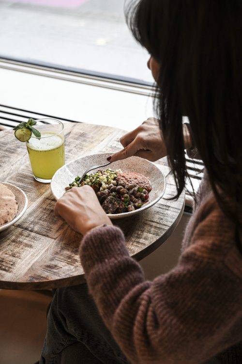 Personne en train de manger un bowl méditerranéen avec une boisson verte sur une table en bois près d’une fenêtre.