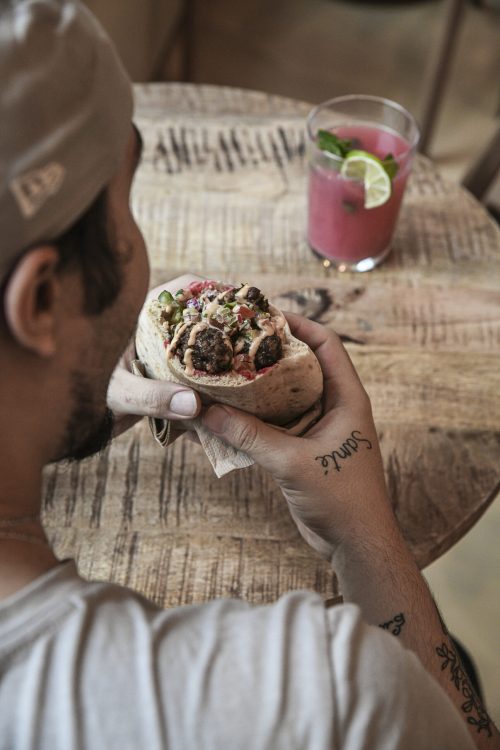 Client dégustant une pita Dip Honest Food bien garnie, avec un cocktail rose sur une table en bois.